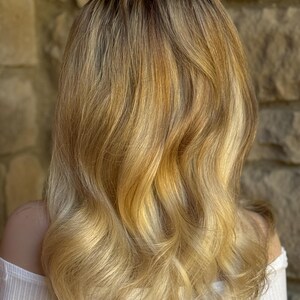 May include: Shoulder-length wavy blonde hair with darker roots and golden highlights. The hair is styled with soft waves and a slight ombre effect, creating a natural and sun-kissed appearance. The person is wearing a white off-the-shoulder top.