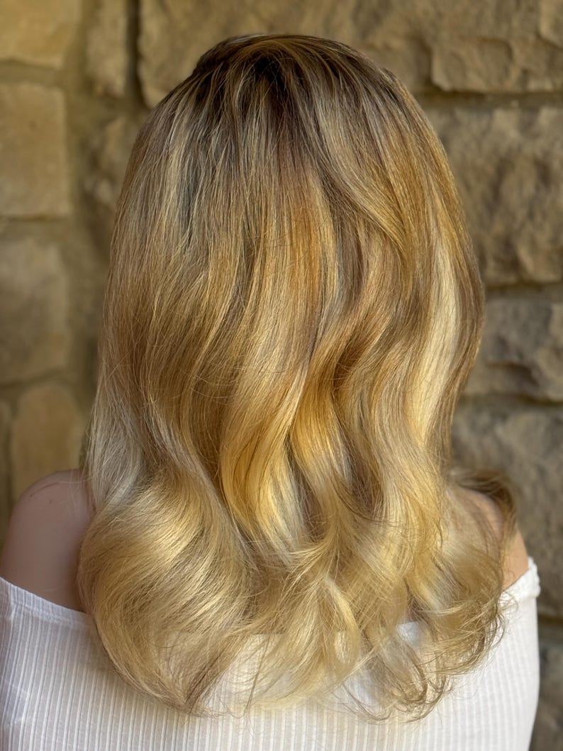 May include: Shoulder-length wavy blonde hair with darker roots and golden highlights. The hair is styled with soft waves and a slight ombre effect, creating a natural and sun-kissed appearance. The person is wearing a white off-the-shoulder top.