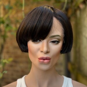 May include: A mannequin head with a short, dark brown bob wig. The wig has a side part and bangs that frame the face. The mannequin is wearing a white tank top and has makeup applied, including lipstick and eyeshadow.