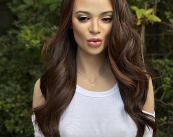 May include: A mannequin head with a brown wig featuring long, wavy hair with highlights. The mannequin is wearing a white, off-the-shoulder ribbed tank top and a gold necklace with a small pendant. The background is a blurred view of green foliage.