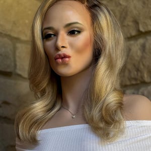 May include: A mannequin head with long, wavy blonde hair, styled with a side part. The mannequin is wearing a white off-the-shoulder top and a delicate silver necklace with a small pendant. The background is a textured stone wall.