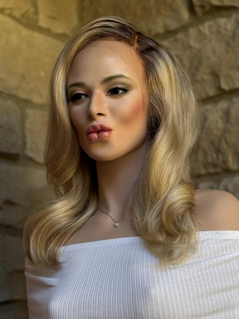May include: A mannequin head with long, wavy blonde hair, styled with a side part. The mannequin is wearing a white off-the-shoulder top and a delicate silver necklace with a small pendant. The background is a textured stone wall.