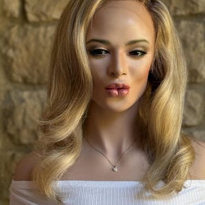 May include: A mannequin head with a blonde wig featuring wavy layers and dark roots. The mannequin is wearing a white off-the-shoulder top and a delicate gold necklace with a small pendant. The background is a stone wall.