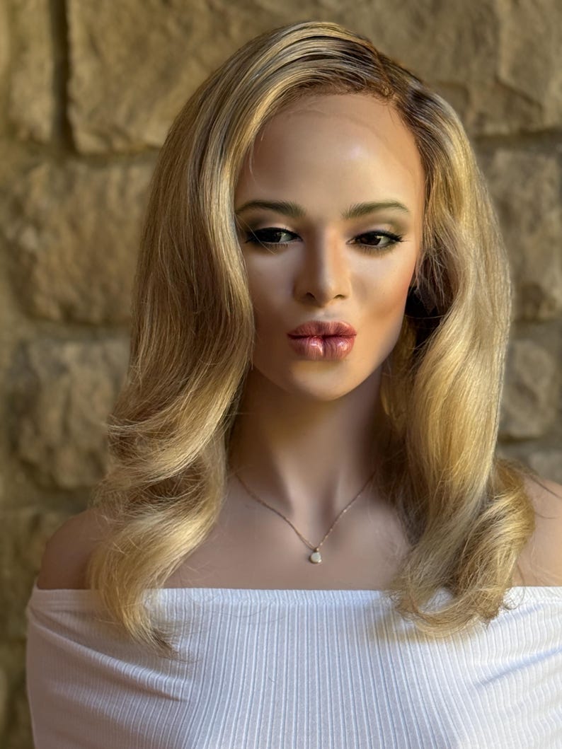 May include: A mannequin head with a blonde wig featuring wavy layers and dark roots. The mannequin is wearing a white off-the-shoulder top and a delicate gold necklace with a small pendant. The background is a stone wall.