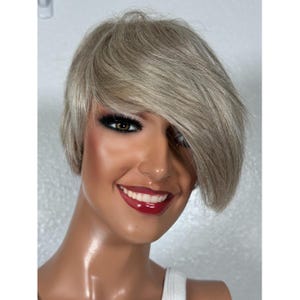 May include: A short, light blonde wig with a side part and bangs. The wig is styled in a modern, edgy look.