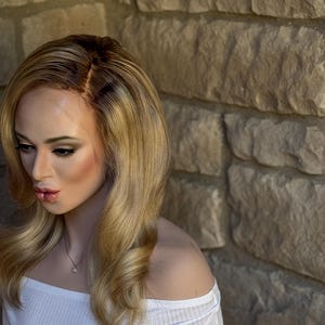 May include: A blonde wig with dark roots and long, wavy layers. The wig is styled with a side part and features a natural-looking hairline. The mannequin is wearing a white off-the-shoulder top and a delicate necklace.