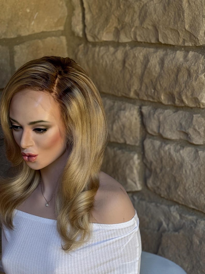 May include: A blonde wig with dark roots and long, wavy layers. The wig is styled with a side part and features a natural-looking hairline. The mannequin is wearing a white off-the-shoulder top and a delicate necklace.