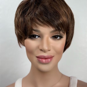 May include: A short, layered brown wig with a side-swept bang. The wig is styled to frame the face and has a natural-looking texture. The person is wearing a white tank top and a gold necklace with a small pendant.