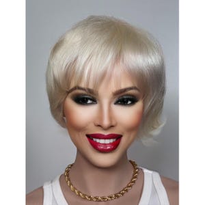 Puede incluir: Una mujer con el pelo corto y rubio platino, lleva un collar de cadena de oro y un top blanco sin mangas. Lleva maquillaje de ojos oscuros y pintalabios rojo.