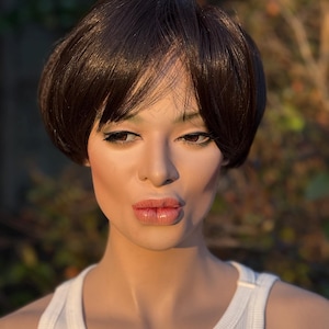 May include: A mannequin wearing a short, dark brown wig with a side part. The mannequin is wearing a white ribbed tank top. The background is blurred with green and brown tones.