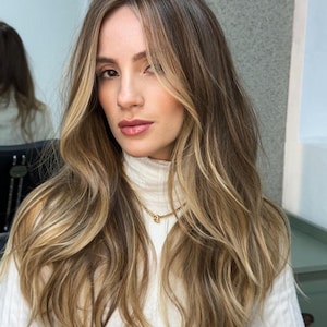 May include: A woman with long, wavy hair featuring a blend of brown, blonde, and light highlights. She wears a white turtleneck sweater and a gold necklace. The hair is styled with a side part and face-framing layers.