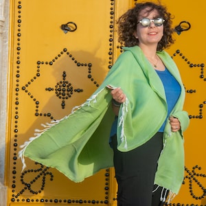 May include: A woman wearing sunglasses and a green shawl with white fringe stands in front of a yellow door with black accents.