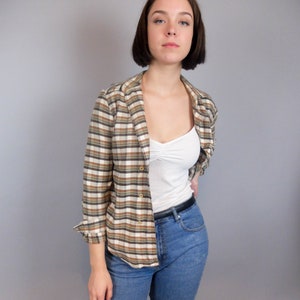 May include: A woman wearing a brown and white checked shirt with a white vest top underneath. She is also wearing blue jeans and a black belt.