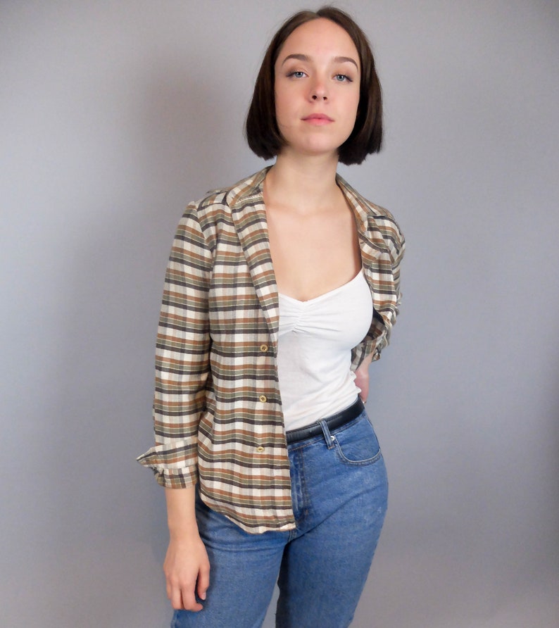 May include: A woman wearing a brown and white checked shirt with a white vest top underneath. She is also wearing blue jeans and a black belt.