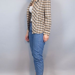 May include: A woman wearing a brown and white checked shirt with a white undershirt, blue jeans, and white trainers.