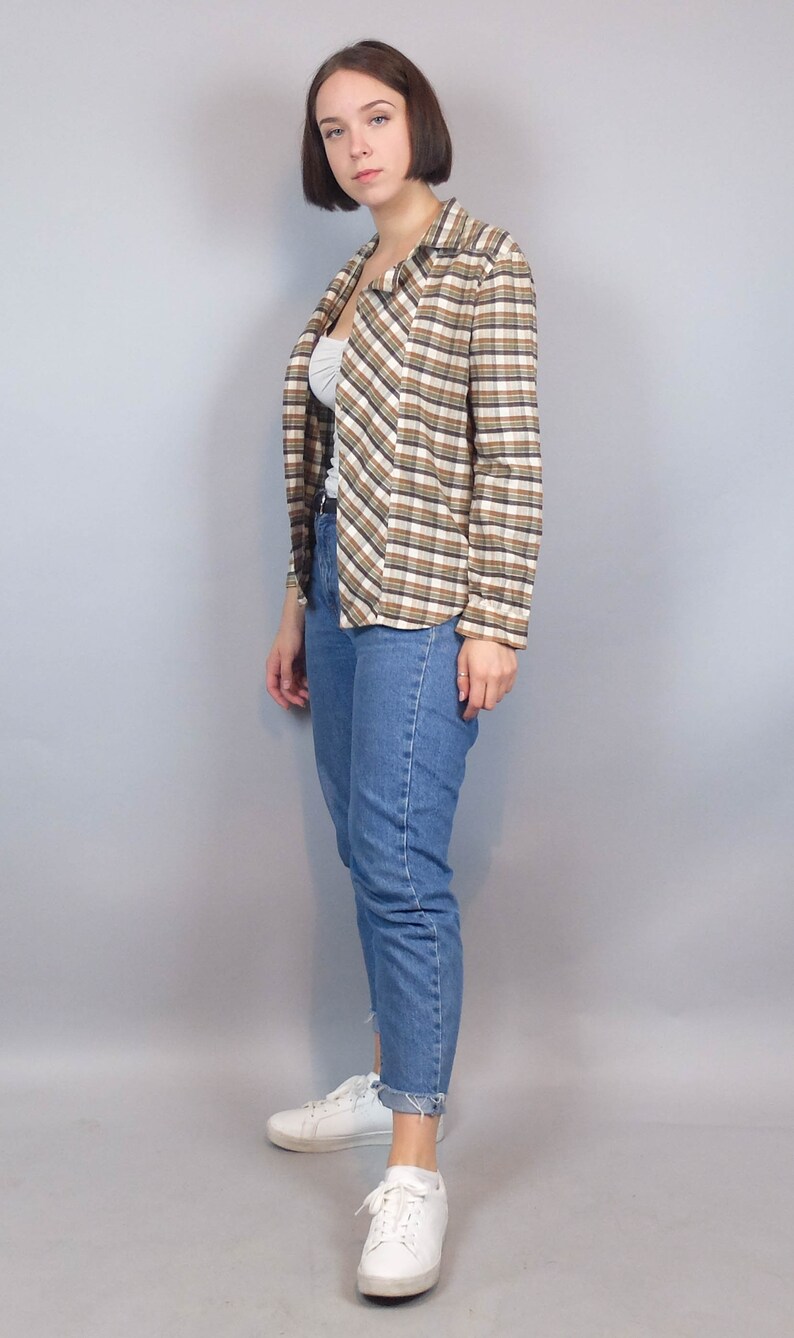 May include: A woman wearing a brown and white checked shirt with a white undershirt, blue jeans, and white trainers.