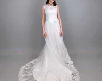 May include: A floor-length white gown featuring a sleeveless lace bodice with a high neckline, a fitted waist accented by a fabric flower, and a flowing tulle skirt. The hem and train are decorated with intricate white floral lace embroidery.