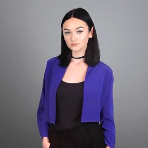 May include: A cropped, royal blue blazer with long sleeves. The blazer is worn over a black top and black pants. The background is a solid gray.
