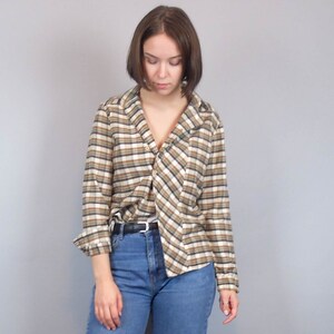 May include: A woman wearing a brown and white checked shirt with a long sleeve and a collared neckline. The shirt is undone and the collar is turned down. The woman is wearing blue jeans and has short brown hair.