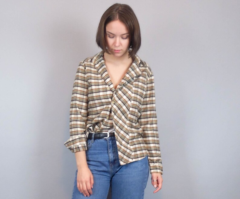 May include: A woman wearing a brown and white checked shirt with a long sleeve and a collared neckline. The shirt is undone and the collar is turned down. The woman is wearing blue jeans and has short brown hair.
