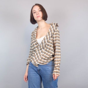 May include: A woman wearing a brown and white checked shirt with a white vest top underneath. She is wearing blue jeans and has her hair tied back.
