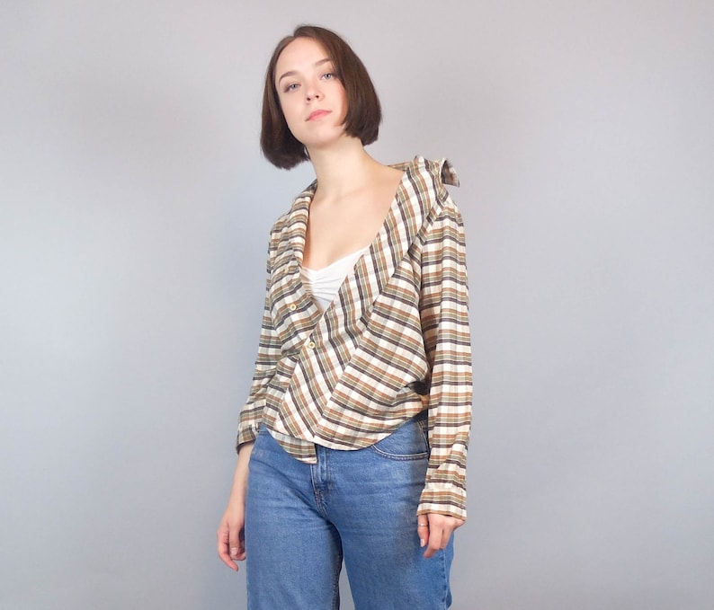 May include: A woman wearing a brown and white checked shirt with a white vest top underneath. She is wearing blue jeans and has her hair tied back.