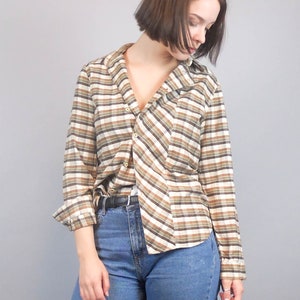 May include: A long-sleeved plaid shirt with brown and white stripes. The shirt is buttoned up and has a collar. The person wearing the shirt is also wearing blue jeans.
