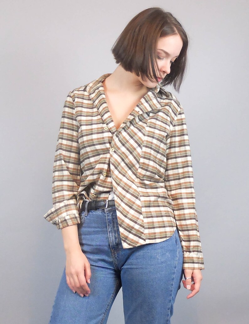 May include: A long-sleeved plaid shirt with brown and white stripes. The shirt is buttoned up and has a collar. The person wearing the shirt is also wearing blue jeans.