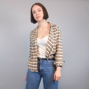 May include: A woman wearing a brown and white checked shirt over a white vest top and blue jeans. She is wearing a black belt.