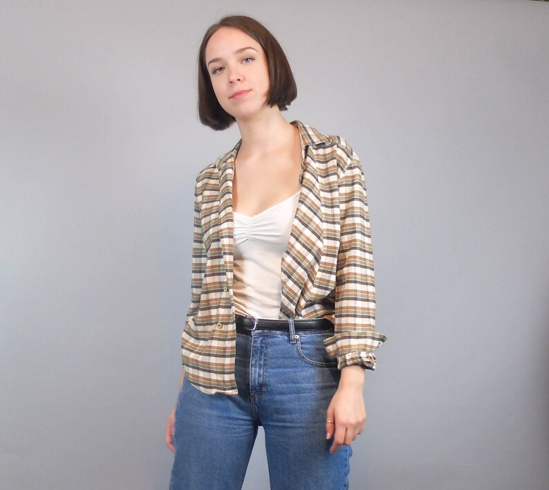 May include: A woman wearing a brown and white checked shirt over a white vest top and blue jeans. She is wearing a black belt.