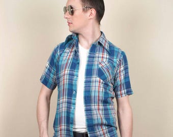 May include: A blue, white, and red plaid short-sleeved shirt with a collared neckline and a chest pocket. The shirt is worn over a white t-shirt, with black trousers and a black belt. The shirt's pattern is a classic design.
