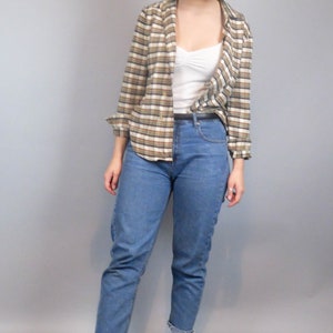 May include: A woman wearing a brown and white checked shirt over a white vest top, blue jeans, and white trainers.