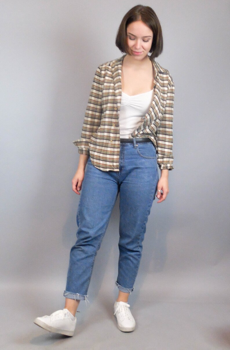 May include: A woman wearing a brown and white checked shirt over a white vest top, blue jeans, and white trainers.