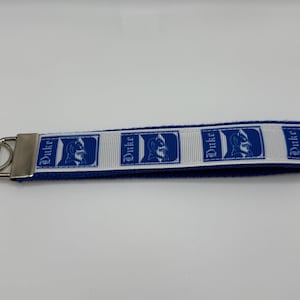 May include: A blue and white keychain wristlet featuring the Duke University logo. The wristlet has a blue base with a white ribbon displaying the Duke logo and the word "Duke" in blue. A silver metal key ring is attached.