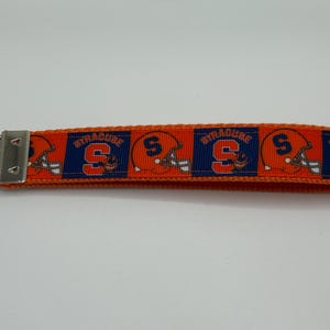 May include: Orange and blue key fob featuring a repeating pattern of Syracuse University football helmets and the word "SYRACUSE". The key fob has a silver metal clasp and key ring.