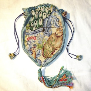 May include: A tapestry drawstring pouch with a detailed design featuring a rabbit, flowers, and a bird. The pouch has a light blue velvet trim and drawstring, with a colourful tassel. The interior fabric has a floral and animal pattern.