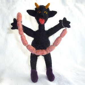 May include: A black felt toy devil with horns and a red mouth holding a string of pink felt sausages.