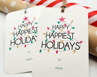 Może przedstawiać: Dwie białe etykiety na prezenty z czarnym napisem "Happy Happiest Holidays", z łańcuchem kolorowych światełek w kształcie choinki. Każda etykieta ma nadrukowane "To:" i "From:". Rolki papieru do pakowania w tle.