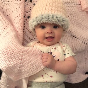 May include: A baby wearing a pink crocheted hat with a faux fur trim and a white onesie with pink and purple paint splatters. The baby is sitting on a pink crocheted blanket.