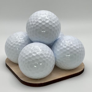 May include: A stack of five white golf balls with dimpled surfaces, resting on a small, square wooden coaster. The golf balls are arranged in a pyramid shape, showcasing their spherical form.