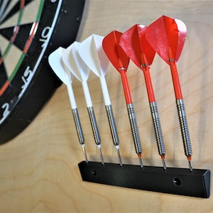 May include: A black dartboard with white and green numbers, and a black dart holder with six darts, three red and three white, mounted on a light wood wall.