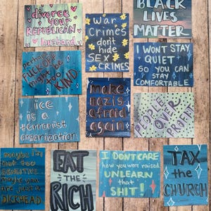May include: A collection of hand-painted wooden signs with various phrases and statements in different colors. The signs include messages like "Black Lives Matter," "Eat the Rich," and other political and social commentary.