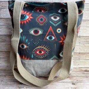 May include: A tote bag with a dark blue background featuring a repeating pattern of stylized eyes in red, orange, and teal. The bag has tan straps and a gray bottom section. The bag is made of fabric.
