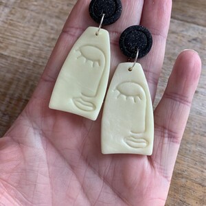 Könnte beinhalten: Ein Paar handgefertigte Ohrringe mit minimalistischem Gesichtsdesign. Die Ohrringe haben eine cremefarbene, längliche Form mit einem schwarzen, kreisförmigen Stift oben. Das Gesichtsdesign umfasst ein geschlossenes Auge und einen einfachen Mund.