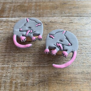 May include: Two gray ceramic opossum earrings with pink details. The opossums are depicted in a curled up position with their tails curled around their bodies.