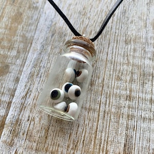 May include: A glass bottle necklace with a cork stopper filled with small white eyeballs. The necklace is made with a black leather cord.