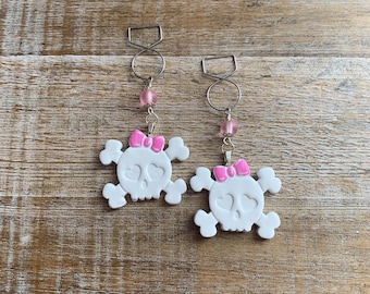 May include: A pair of white skull earrings with pink bows and pink beads. The earrings are hanging from silver hoops.