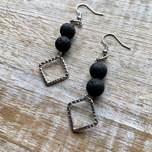 May include: A pair of dangle earrings featuring black lava beads and silver-toned, hammered-look square charms. The earrings have a simple design with a hook closure. The background is a light-colored, wooden surface.