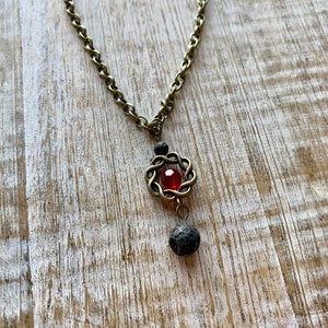 May include: A bronze chain necklace with a red gemstone and black lava rock beads. The pendant features a circular design with a woven pattern.