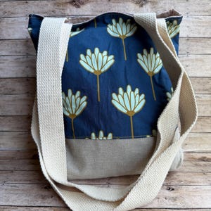 Puede incluir: Un bolso tote azul marino con un estampado floral en azul claro y dorado. El bolso tiene una base beige y correas color crema. El bolso está hecho de tela y está diseñado para transportar artículos.